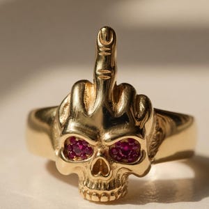 Op de afbeelding: Een goudkleurige ring met een schedelontwerp en robijnrode edelstenen als ogen. De middelvinger van de schedel is omhoog. De ring is een opvallend sieraad voor liefhebbers van unieke sieraden.