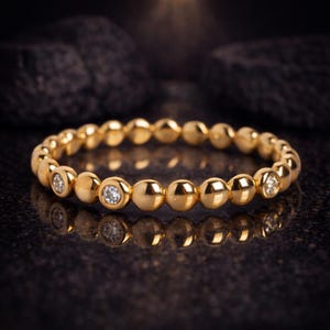 Könnte beinhalten: Ein goldener Ring mit einer Reihe kleiner, runder Perlen. Der Ring hat mehrere klare, runde Edelsteine, die in das Design eingesetzt sind. Der Ring wird auf einer dunklen, reflektierenden Oberfläche präsentiert.