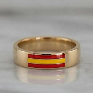 Puede incluir: Un anillo de banda de metal dorado con una incrustación horizontal de tres franjas de esmalte en rojo, amarillo y rojo. El anillo tiene un acabado liso y pulido con un perfil plano, mostrando un diseño tricolor en la superficie exterior.