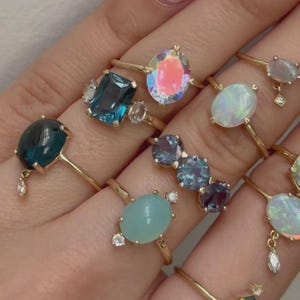 May include: A collection of gold rings with various gemstones, including blue, teal, and iridescent stones. The rings have delicate gold bands and are set with oval, pear, and rectangular cut gems. Some rings feature small accent stones.