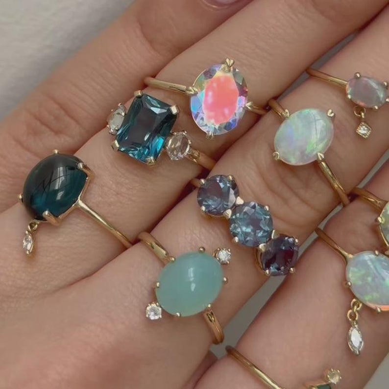 May include: A collection of gold rings with various gemstones, including blue, teal, and iridescent stones. The rings have delicate gold bands and are set with oval, pear, and rectangular cut gems. Some rings feature small accent stones.