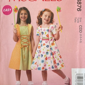 May include: A sewing pattern for two girls' dresses. The pattern is called McCall's M6876 and is for sizes 2-3-4-5. The dresses are both A-line and have a floral print. One dress is yellow and orange with white polka dots and the other is white with a multi-colored floral print. The pattern is labeled "Easy" and is part of the "Ruffles and Lace Treasured Collection".