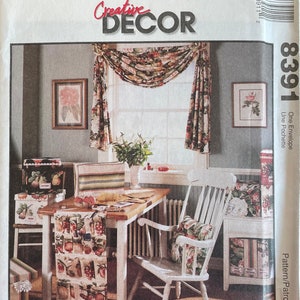 May include: A McCall's Creative Decor sewing pattern for a window treatment with a floral print. The pattern number is 8391. The image shows a room with a window, a table, and chairs. The window treatment is a valance with a swag and a floral print. The table is set with a runner and a floral print. The chairs are white with floral print cushions.