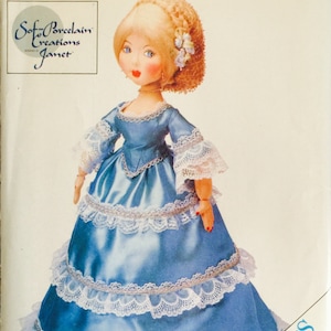 May include: A vintage sewing pattern for a porcelain doll wearing a blue dress with white lace trim. The pattern is titled "Soft Porcelain Creations Janet" and is from Butterick Craft.