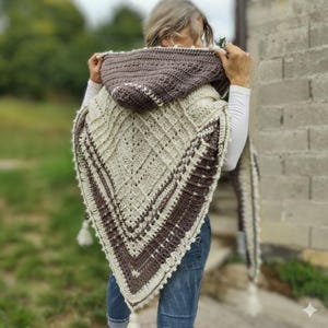 Peut inclure: Un châle à capuche en crochet crème et marron. Le châle présente un motif géométrique avec une bordure et une capuche marron. La capuche est relevée, mettant en valeur le design. Le châle a un aspect texturé et une gland à l'extrémité.