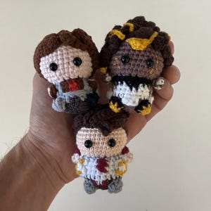 May include: Three small, crocheted dolls with brown hair and black eyes. The dolls have different outfits, including a gray and red outfit, a black and white outfit with yellow accents, and a white and red outfit with yellow accents. The dolls are held in a hand.