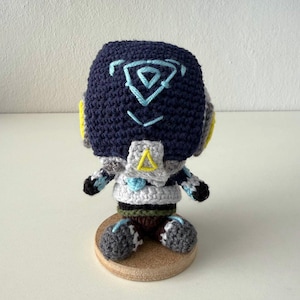 Könnte beinhalten: Gehäkelte Puppe mit einem marineblauen Kopf mit hellblauem geometrischen Design. Die Puppe hat gelbe Akzente, graue und weiße Details und ist auf einem kleinen Holzsockel montiert. Der Körper ist hauptsächlich weiß und grau.