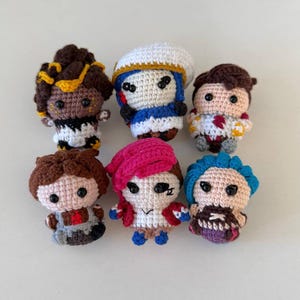 May include: A collection of seven handmade crochet dolls, each with unique features and outfits. The dolls have various hair colors, including brown, pink, and blue, and wear different clothing. The dolls are approximately 3 inches tall.