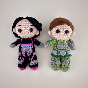 May include: Two crocheted dolls, one in a black and pink outfit, the other in green and gray. Both dolls have black eyes, light skin, and brown or black hair. The dolls are handmade and appear to be characters from a science fiction theme.
