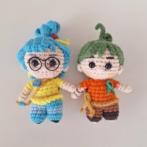 Op de afbeelding: Twee handgemaakte gehaakte poppen die elkaars hand vasthouden. De ene pop heeft blauw haar, een bril, een geel shirt en een blauwe broek. De andere pop heeft groen haar, een oranje shirt en een bruine broek. Beide poppen hebben zwarte knoopogen.