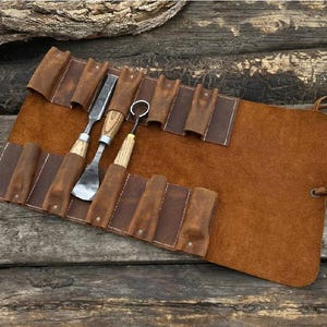 May include: A brown leather tool roll with several wood-handled carving tools. The roll is open, displaying the tools and their storage slots. The leather has a natural, textured appearance, and the tools are made of metal and wood.