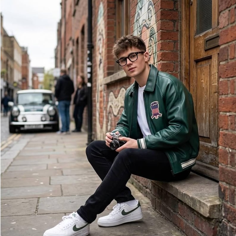 May include: A green leather jacket with a white stripe on the cuffs and a patch on the chest. The jacket is worn with a white t-shirt, black jeans, and white sneakers with a green swoosh. A vintage camera is held.