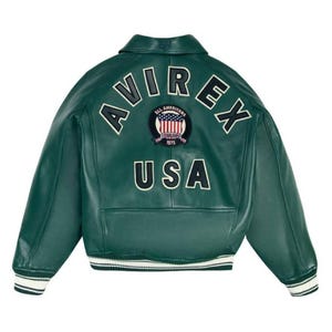 May include: A dark green leather jacket with the word "AVIREX" arched across the upper back in white lettering. Below the lettering is a patch with the words "ALL AMERICANS ESTABLISHED 1975" and a USA flag design. The word "USA" is below the patch.
