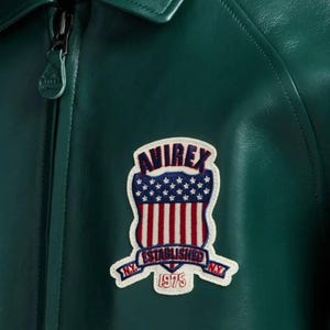 May include: A close-up of a dark green leather jacket with a zipper and a patch. The patch features the word "AVIREX" above a design of the American flag and the words "ESTABLISHED 1975 N.Y."