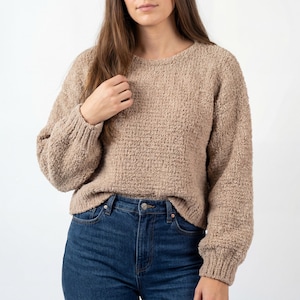 Könnte beinhalten: Ein beigefarbener Pullover mit Rundhalsausschnitt und langen Ärmeln. Der Pullover hat ein strukturiertes Strickdesign und gerippte Bündchen und Saum. Das Model trägt den Pullover mit einer blauen Jeans.