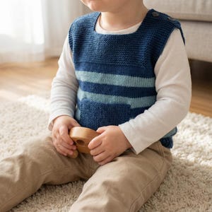 Puede incluir: Un chaleco azul tejido con rayas azul claro y un bolsillo. El chaleco tiene dos botones en el hombro y se usa sobre una camisa blanca de manga larga. El niño sostiene un juguete de madera y lleva pantalones color caqui.