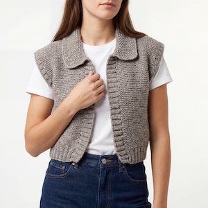 May include: A light brown, knitted vest with a collar. The vest is sleeveless and has a button-up front. The model is wearing a white t-shirt and blue jeans.