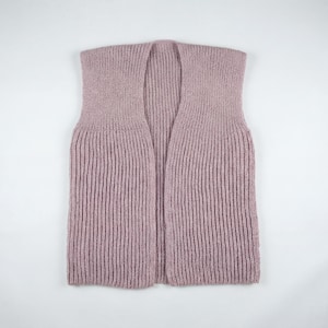 Puede incluir: Un chaleco de punto sin mangas de color malva. El chaleco presenta un diseño acanalado en todo el cuerpo y las mangas. La prenda está colocada sobre una superficie blanca, mostrando su diseño abierto y su textura.
