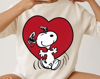 Könnte beinhalten: Cremefarbenes Sweatshirt mit einem großen roten Herz und der Cartoonfigur Snoopy in der Mitte. Snoopy ist weiß mit schwarzen Akzenten und springt vor Freude. Das Sweatshirt hat lange Ärmel.
