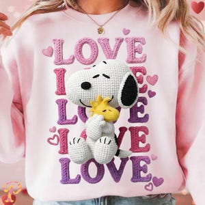 May include: A light pink sweatshirt featuring a crochet Snoopy and Woodstock design. The word "LOVE" is vertically and horizontally displayed in a knitted style, with pink and purple colors. Small pink hearts are scattered around the design.