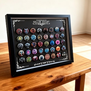 May include: A framed display featuring a collection of colorful, circular pins. Each pin showcases a unique design with intricate details and vibrant hues. The frame is black, and the pins are arranged on a black background, with the text "Hollow Knight" at the top.