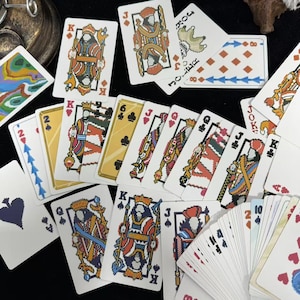 Puede incluir: Un juego de cartas con ilustraciones pixeladas de reyes, reinas y jotas. Las cartas presentan palos de picas, corazones, tréboles y diamantes. Algunas cartas están abanicadas, mostrando sus coloridos diseños y números.