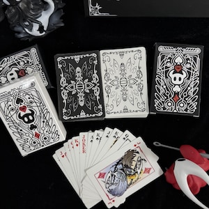 Puede incluir: Un conjunto de naipes y cajas de cartas con intrincados diseños en blanco y negro. Las cartas presentan un motivo de calavera y corazón, con un abanico de cartas ilustradas. Las cajas y cartas están dispuestas sobre una superficie negra.