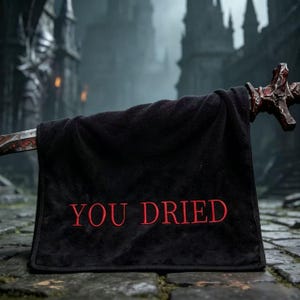 Puede incluir: Toalla negra con el texto bordado "YOU DRIED" en rojo. La toalla está drapeada sobre una espada con una empuñadura detallada. El fondo presenta una estructura de piedra de estilo medieval.