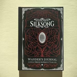 Puede incluir: Un diario negro con el título "Hollow Knight Silksong" en letras blancas. La portada presenta un diseño decorativo rojo y el texto "Wander's Journal: A Journal Through the Heart of Pharloom". El diario tiene un borde blanco.