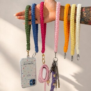 Può includere: Una collezione di sette cordini da polso in macramè nei colori verde oliva, blu, magenta, rosa chiaro, arancione, giallo e crema. Un cordino regge una cover trasparente con la scritta 'iP 12 Mini (5.4)', un altro un astuccio rosa e uno chiavi.