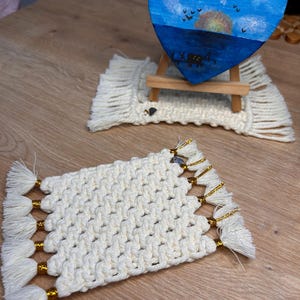 May include: Two rectangular macrame coasters made of off-white cord with gold metallic thread accents and fringed edges. One coaster sits under a small wooden easel holding a heart-shaped painting of a blue sky, sunset, and dark silhouettes.