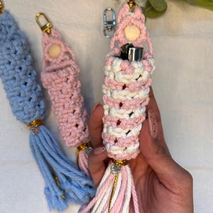 Puede incluir: Soportes para encendedores de macramé hechos a mano en tonos rosa, blanco y azul, con herrajes dorados y borlas. Los soportes están hechos a mano con un diseño tejido, y se ve un encendedor dentro de uno de los soportes rosa y blanco. Un accesorio decorativo.