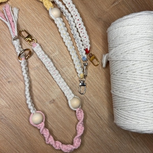 Op de afbeelding: Close-up van handgemaakte macramé lanyards in verschillende kleuren, waaronder wit, roze, geel, rood en blauw. Elke lanyard heeft houten kralen en goudkleurige metalen sluitingen. Een grote spoel wit katoenen koord is zichtbaar.
