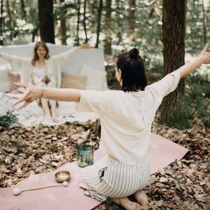 May include: A yoga scene in a forest setting. A person is kneeling on a pink yoga mat, arms outstretched. A singing bowl, mallet, and a teal glass jar are on the mat. Another person is in the background, also with arms outstretched.