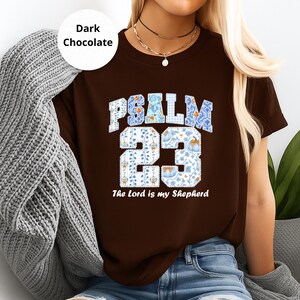 Könnte beinhalten: Dunkelbraunes T-Shirt mit dem Text "PSALM 23" in einem weißen, blauen und floralen Design. Der Satz "The Lord is my Shepherd" ist darunter gedruckt. Das Shirt ist einfarbig und hat kurze Ärmel.