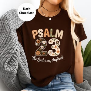 Könnte beinhalten: Dunkelbraunes T-Shirt mit dem Text "PSALM 23" in dekorativer Schriftart, mit floralen Akzenten. Der Satz "The Lord is my shepherd" ist darunter gedruckt. Das Shirt ist Teil einer Bekleidungskollektion.