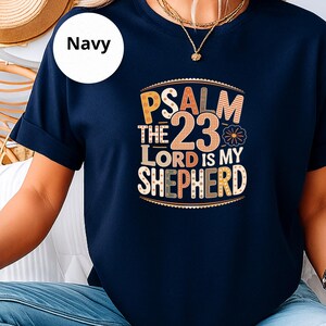 Könnte beinhalten: Marineblaues T-Shirt mit dem Text "Psalm 23 Der Herr ist mein Hirte" in einer farbenfrohen, dekorativen Schriftart. Das Shirt hat kurze Ärmel und einen Rundhalsausschnitt. Das Wort "Navy" befindet sich in einem weißen Kreis oben links.