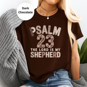 Könnte beinhalten: Dunkelbraunes T-Shirt mit dem Text "PSALM 23 THE LORD IS MY SHEPHERD" in dekorativer Schrift. Das Shirt hat eine einfarbige Farbe mit Rundhalsausschnitt und kurzen Ärmeln. Das Shirt ist dunkelbraun.