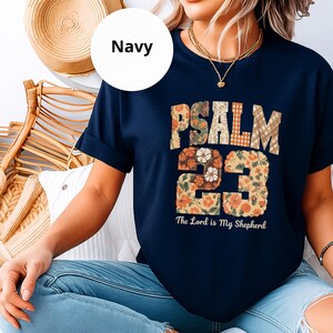 Könnte beinhalten: Marineblaues T-Shirt mit dem Text "PSALM 23" in einem floralen und karierten Design. Der Text "The Lord is My Shepherd" steht unter den Zahlen. Das T-Shirt hat kurze Ärmel.