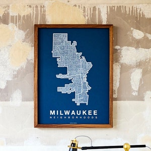 May include: A framed map of Milwaukee, Wisconsin, with a blue background and white lines outlining the city's neighborhoods. The frame is made of wood and has a dark brown finish. The map is titled "Milwaukee Neighborhoods".