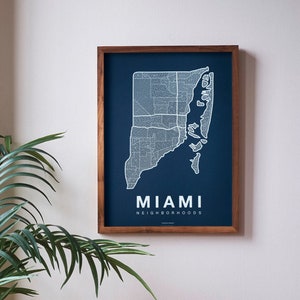 May include: A framed map of Miami neighborhoods with a dark blue background and white lines. The map is hanging on a white wall above a brown leather couch with a patterned pillow.