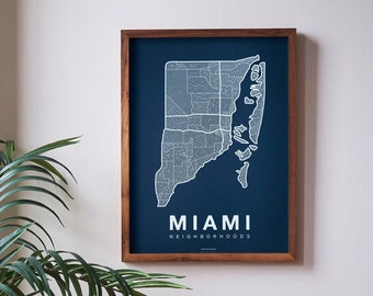 May include: A framed map of Miami neighborhoods with a dark blue background and white lines. The map is hanging on a white wall above a brown leather couch with a patterned pillow.