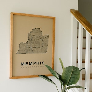 Könnte beinhalten: Eine gerahmte Karte von Memphis, Tennessee, mit dem Titel "Memphis Neighborhoods" unter der Karte. Die Karte hängt an einer weißen Wand neben einer Holztreppe. Eine Topfpflanze steht auf einem Holztisch mit einem Stapel Bücher und einer Glasflasche mit Streichhölzern.