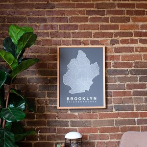 May include: A framed map of Brooklyn neighborhoods hangs on a brick wall. A small table with a lamp and books sits in front of a brown armchair with a blue pillow.