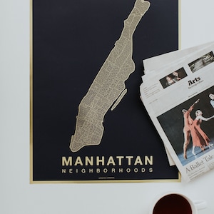 May include: A gold foil map of Manhattan neighborhoods on a dark blue background. The map is framed with a gold border. The text "MANHATTAN NEIGHBORHOODS" is printed in gold at the bottom of the map.