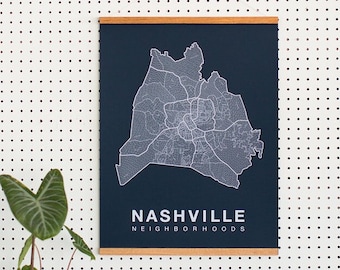 May include: A black and white map print of Nashville neighborhoods on a dark blue background with a wooden frame. The print is hanging on a white pegboard wall with a potted plant in front of it.