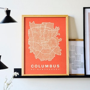 May include: A framed print of a map of Columbus, Ohio, with the city's neighborhoods outlined in white on a bright orange background. The frame is made of wood and has a light brown finish. The print is hanging on a white wall above a couch with orange and gray throw pillows.