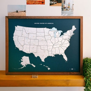 May include: A dark blue framed map of the United States with the text "United States of America" at the top. The map is divided into states and has a white background. The map is on a wooden surface.