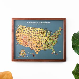 May include: A framed map titled Natural Wonders of the United States of America, featuring a yellow-to-green gradient map of the U.S. on a blue background, decorated with numerous small, colourful illustrations of landmarks and natural sites.