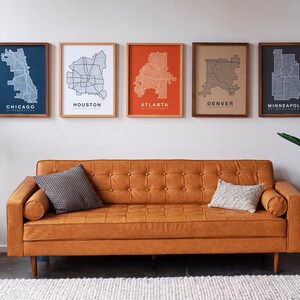 May include: Five framed city maps of Chicago, Houston, Atlanta, Denver, and Minneapolis hang on a white wall above a brown leather sofa with two throw pillows. A potted plant is on the right side of the image and a small wicker table is on the left.